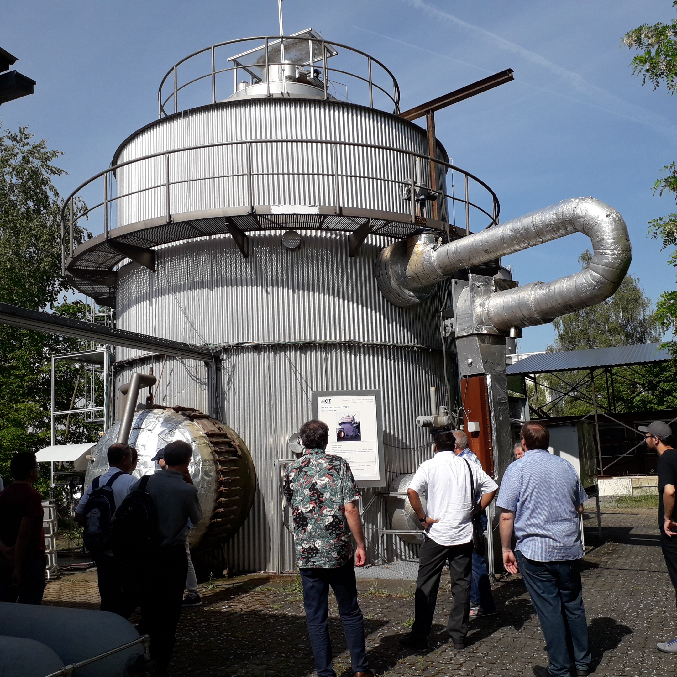 Gruppe vor einer großen, runden Industrieanlage bei sonnigem Wetter.